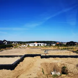 Przygotowanie fundamentów pod budowę domu jednorodzinnego; widoczne zbrojenia, izolacja pionowa i zagęszczarka wibracyjna na placu budowy, w tle zabudowa mieszkaniowa.