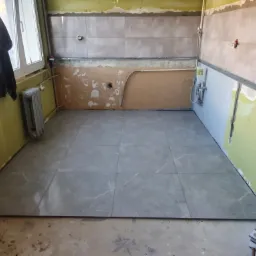 Wnętrze kuchni w trakcie remontu: ułożona szara podłoga z płytek imitujących marmur, częściowo wykończone ściany z widocznymi instalacjami i listwami montażowymi, okno z wiszącą kurtką.