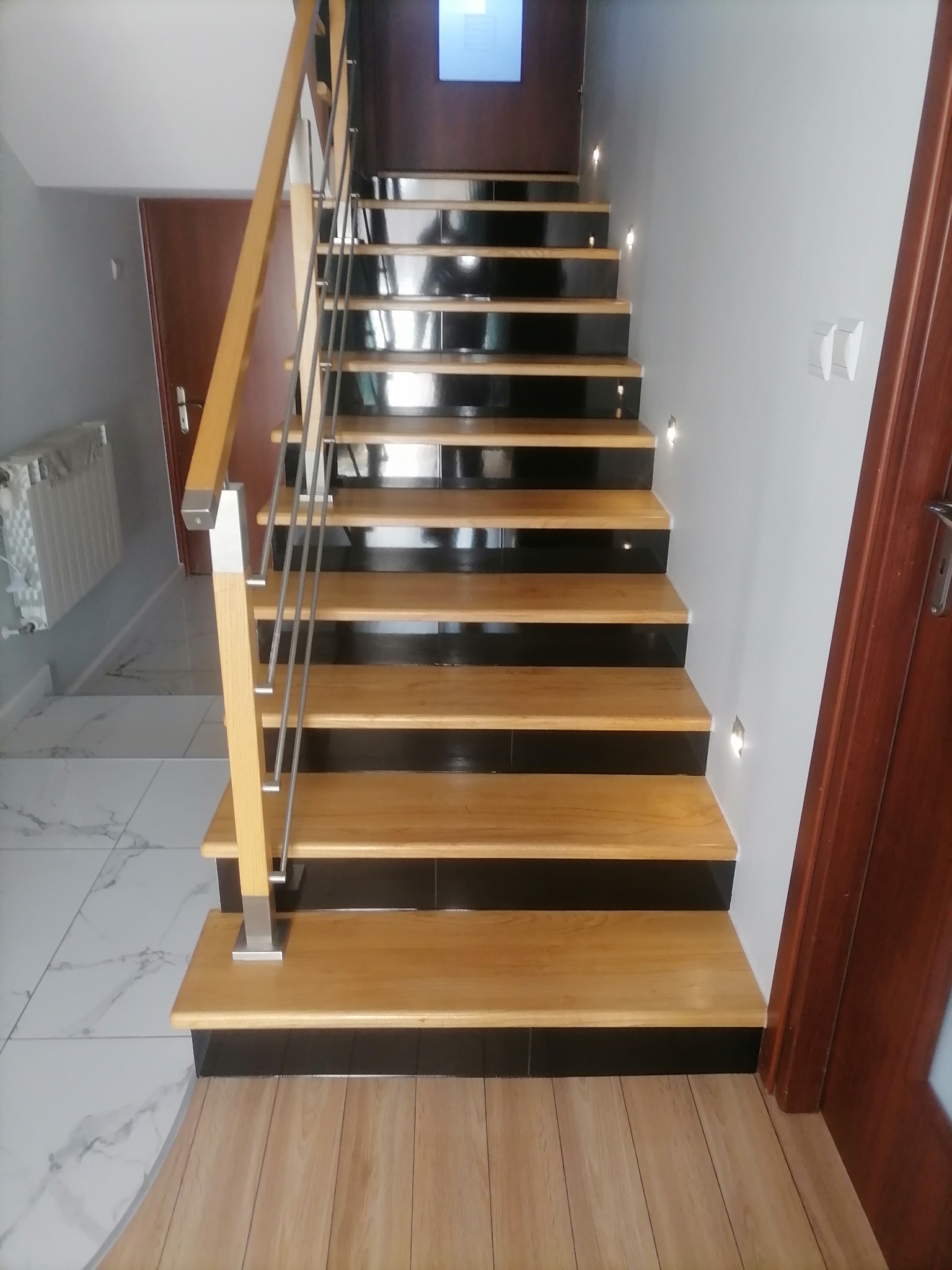 Schody z drewnianymi stopniami i czarnymi podstopnicami, oświetlone punktowymi lampkami w ścianie, z metalową balustradą i drewnianą poręczą, widoczne drzwi w tle.