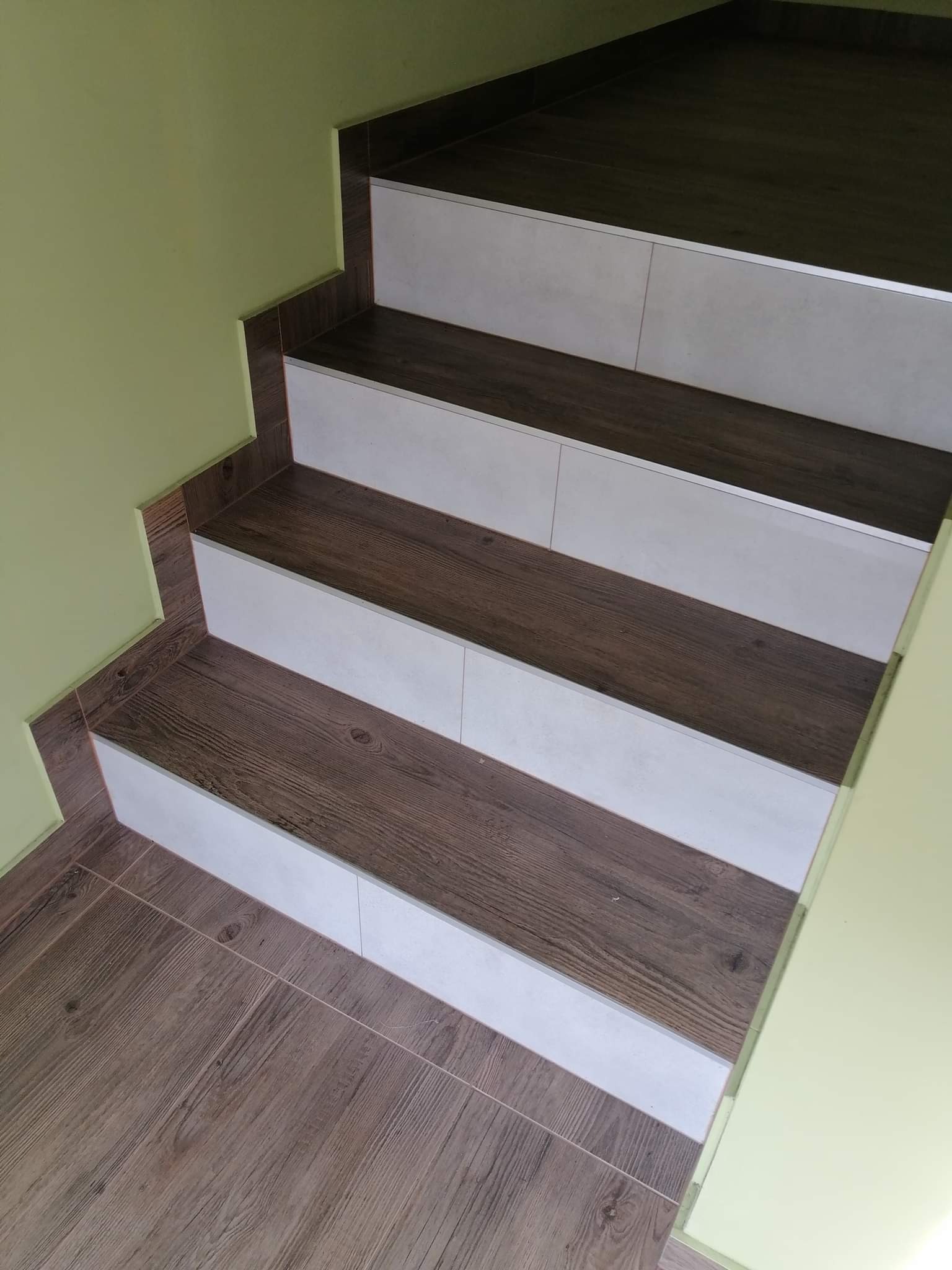 Wykończone schody z płytek imitujących drewno, z białymi wstawkami, prowadzące w górę, przy jasnej, zielonej ścianie.