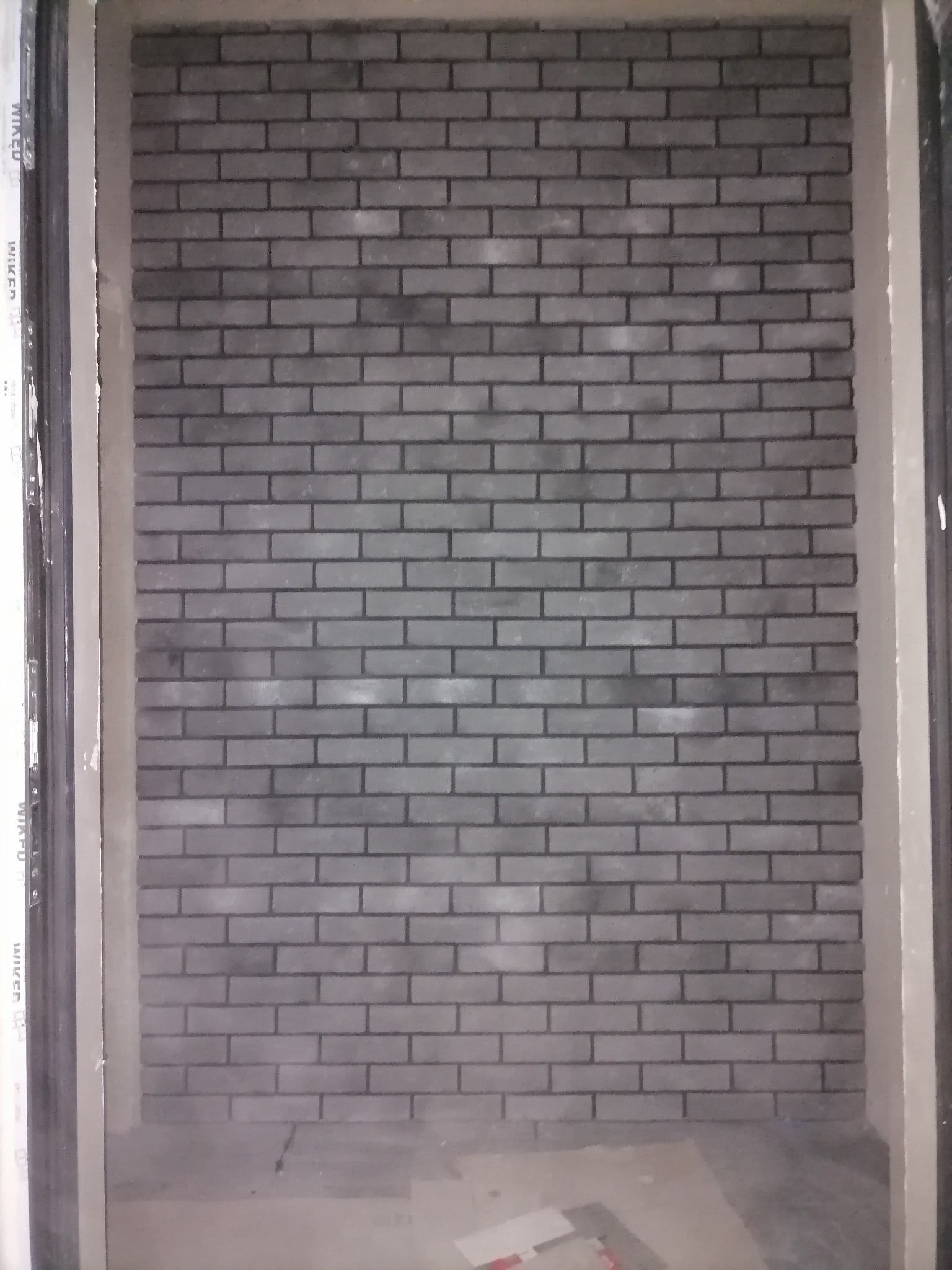 Ściana wykończona szarą cegłą klinkierową, widoczna zza ościeżnicy drzwiowej, z fragmentami płytek na podłodze.