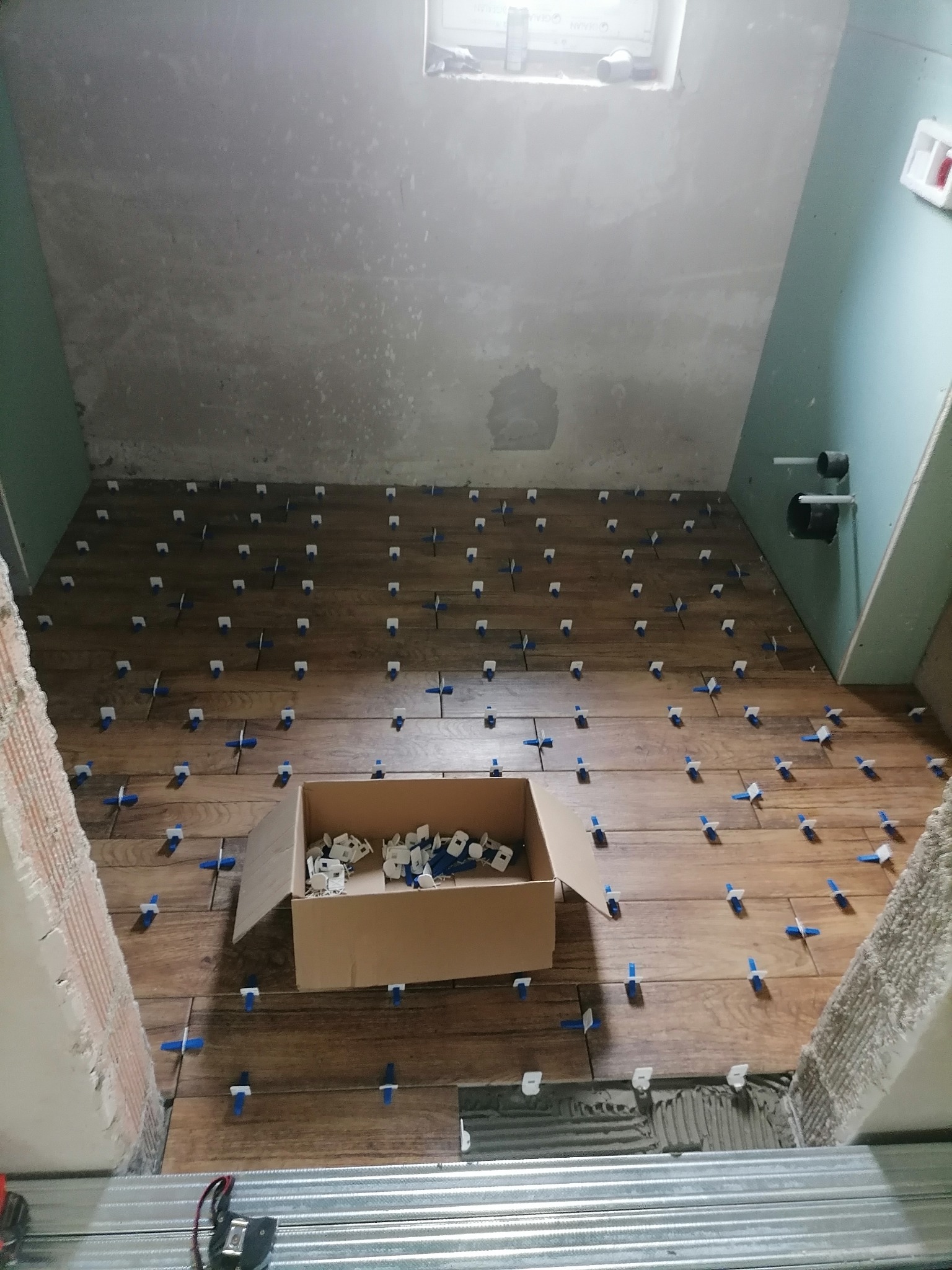 Układanie płytek imitujących drewno na podłodze w łazience z użyciem niebieskich krzyżyków dystansowych i systemu poziomowania płytek, widoczna kartonowa skrzynka z zapasem krzyżyków.