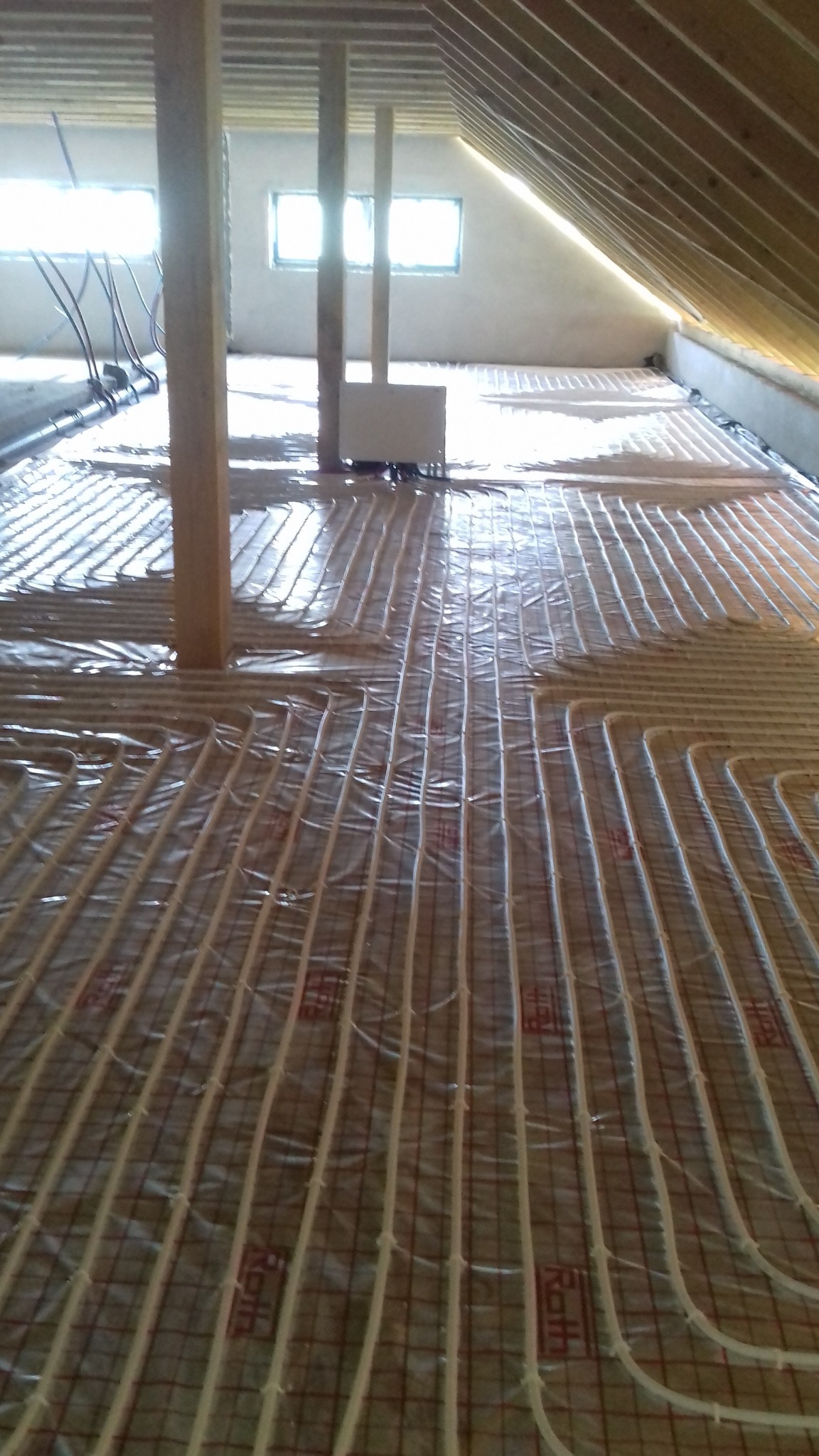 Montaż ogrzewania podłogowego w nowym budynku, widoczne rury grzewcze ułożone na folii z siatką, drewniane słupy podtrzymujące konstrukcję dachu, okna w tle.