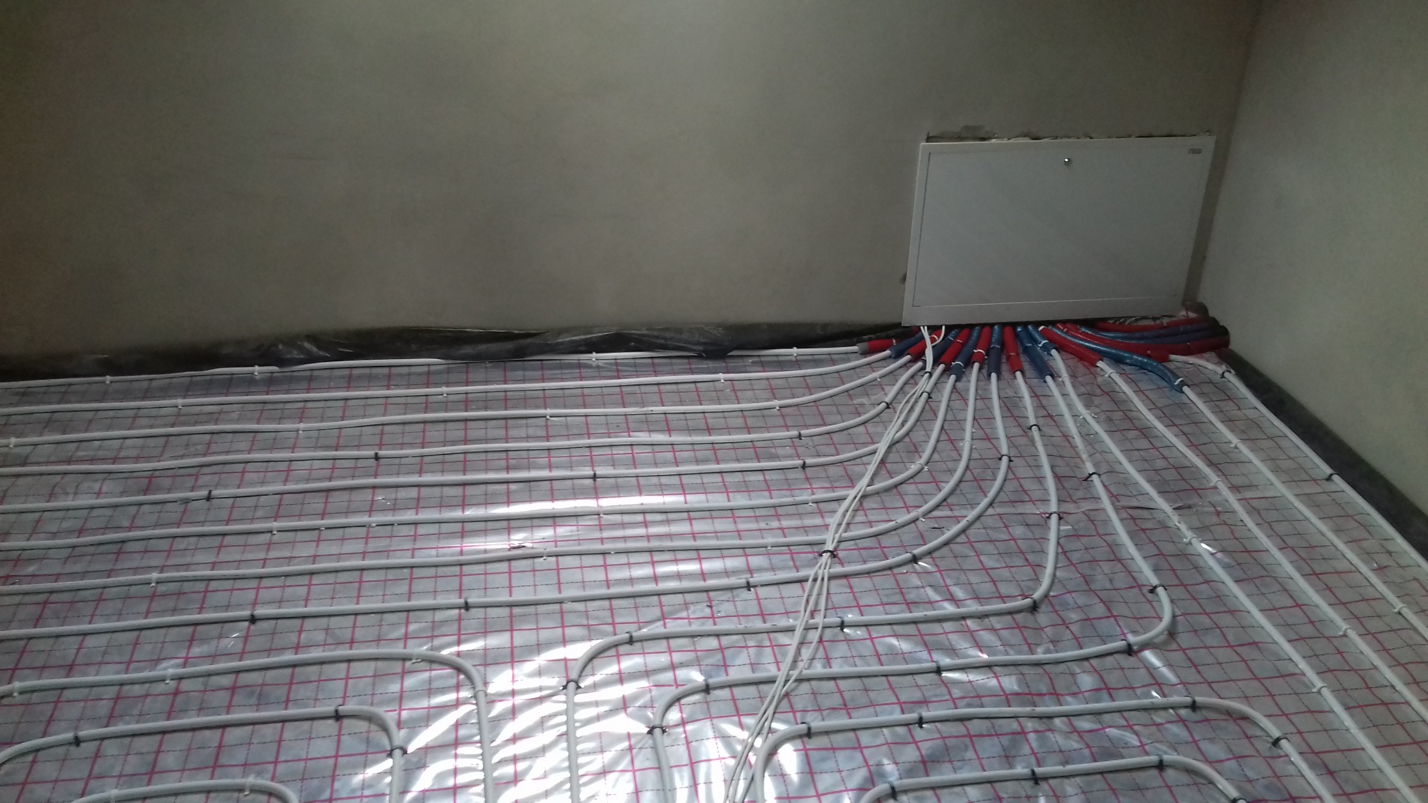 Rozłożona instalacja ogrzewania podłogowego na folii z siatką, z rurami w kolorach czerwonym i niebieskim wchodzącymi do białej skrzynki rozdzielczej przy ścianie.