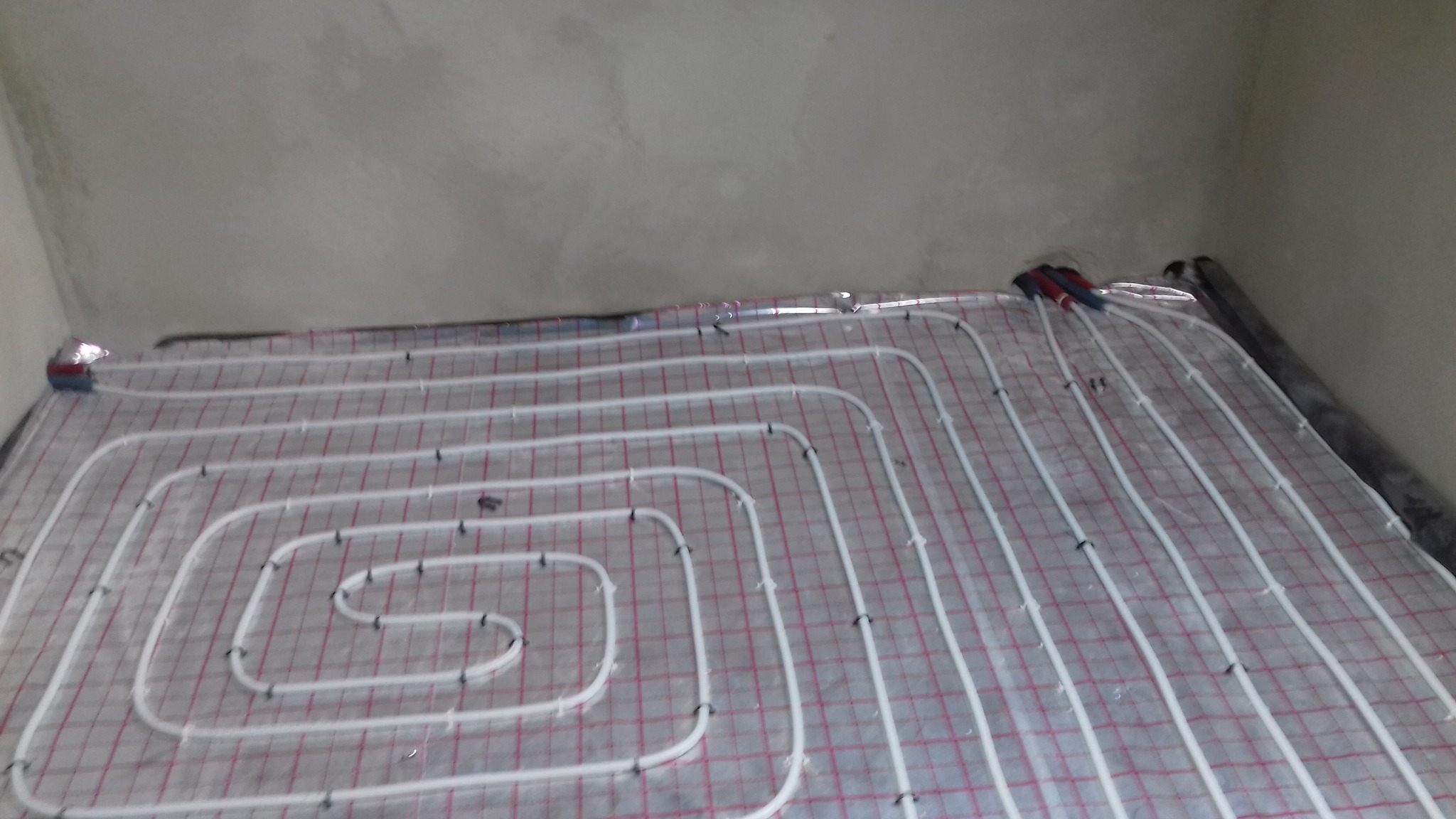 Białe rury ogrzewania podłogowego ułożone spiralnie na folii z czerwoną kratką, zamocowane czarnymi klipsami do podłoża. Widoczny fragment ściany w tle.