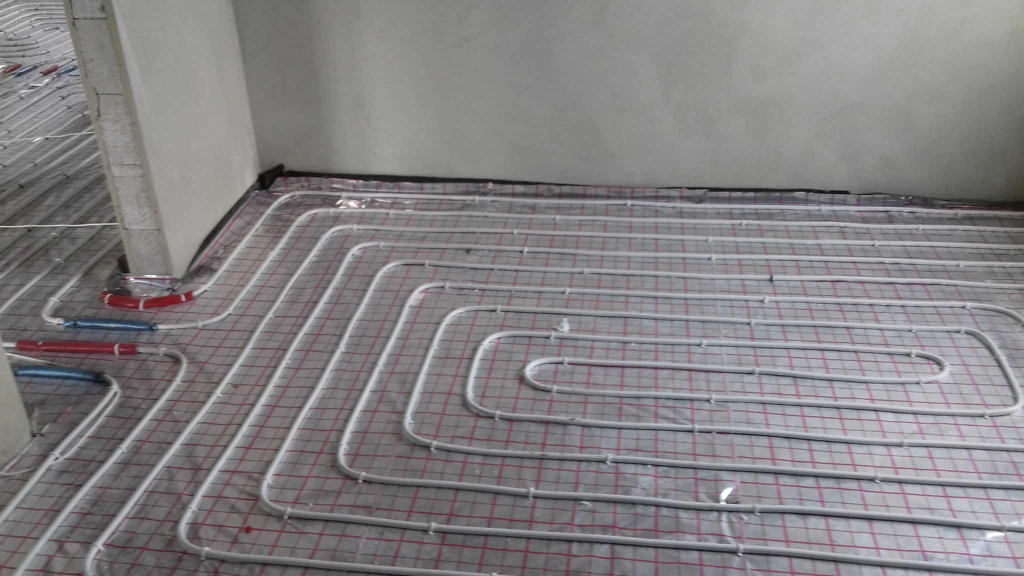 Białe rury ogrzewania podłogowego ułożone spiralnie na siatce z folią aluminiową w surowym wnętrzu, widoczne złącza rur w kolorze czerwonym i niebieskim przy kolumnie