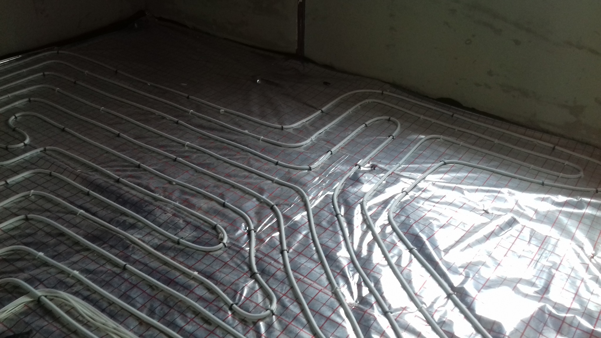 Układ rur ogrzewania podłogowego na folii izolacyjnej z siatką, widok z góry w surowym wnętrzu, rury mocowane plastikowymi klipsami.