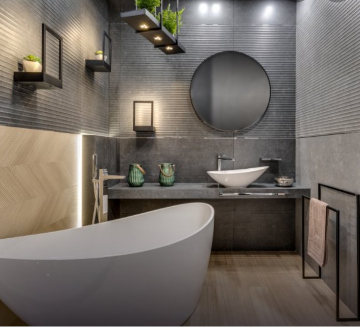 Nowoczesna łazienka z wolnostojącą wanną, okrągłym lustrem i szarymi płytkami z poziomym wzorem, uzupełniona o minimalistyczne półki i rośliny doniczkowe.