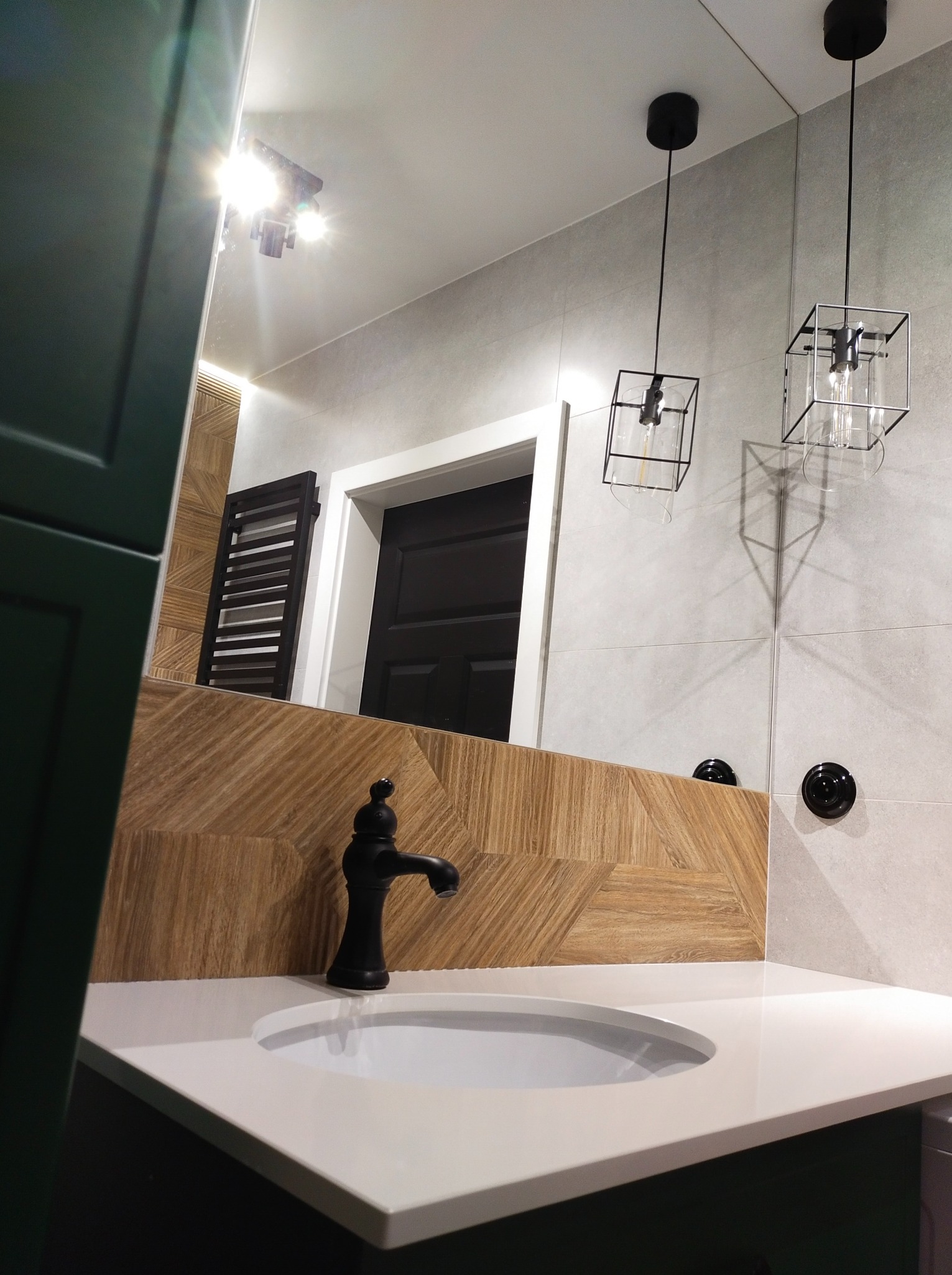 Nowoczesna łazienka z umywalką nablatową, czarną armaturą i geometrycznymi lampami wiszącymi, odbijająca się w dużym lustrze z widocznym grzejnikiem drabinkowym i fragmentem drzwi.