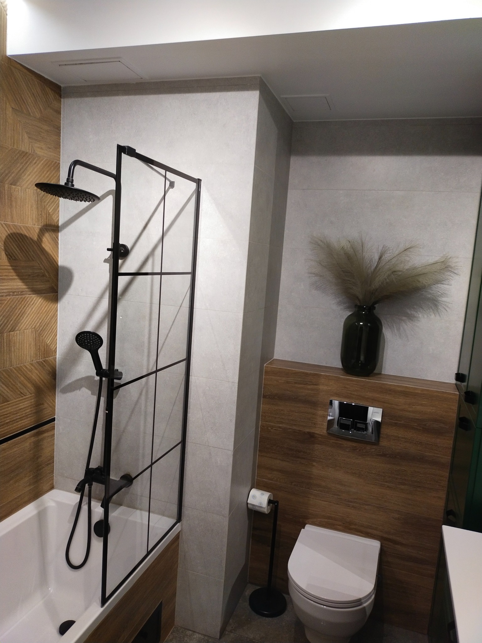 Nowoczesna łazienka z wanną, czarną armaturą natryskową i szklanym panelem prysznicowym, dekorowana drewnopodobną okładziną i wazonem z suszonymi trawami.