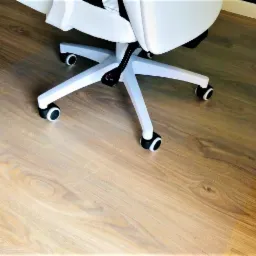 Przezroczysta mata ochronna pod krzesło biurowe na drewnianej podłodze, widoczne kółka białego krzesła i fragment biurka.