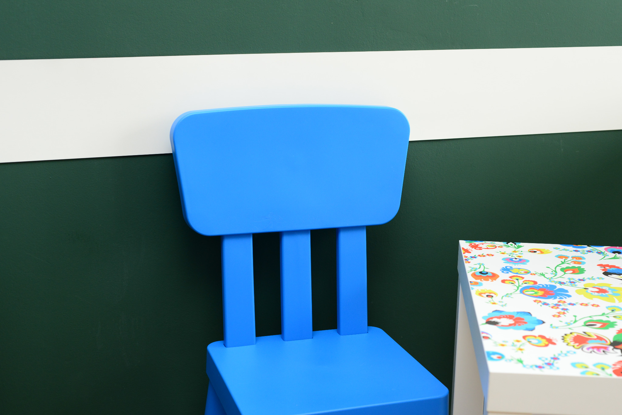 Niebieskie krzesło dziecięce z trzema szczebelkami na oparciu, ustawione tyłem na tle ciemnozielonej ściany z białym pasem. Obok widoczny fragment stolika z blatem w kolorowe kwiaty.