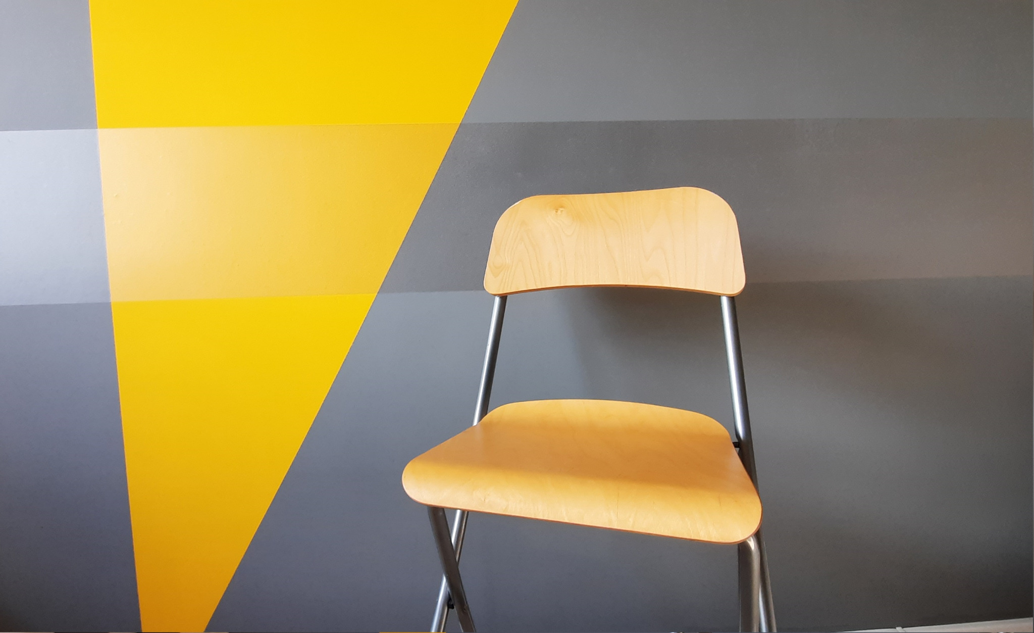 Jasne, składane krzesło z drewnianym siedziskiem i oparciem na tle ściany pomalowanej w geometryczne wzory w odcieniach żółci i szarości.