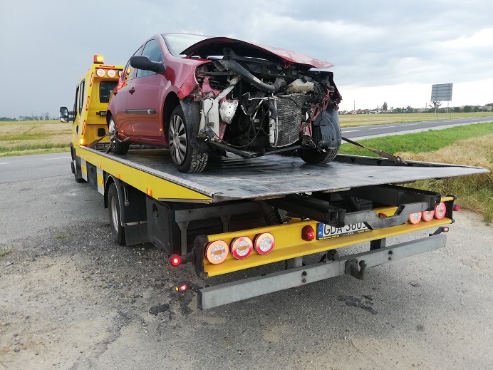 Czerwony uszkodzony samochód osobowy załadowany na żółtą platformę pojazdu pomocy drogowej z widocznymi światłami ostrzegawczymi, na tle polnej drogi i pochmurnego nieba.