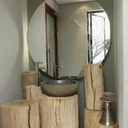 Łazienka z umywalką w kształcie misy, osadzoną na drewnianych pniakach o różnej wysokości, z okrągłym lustrem z delikatnym motywem roślinnym.