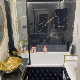 Luksusowa łazienka z czarnymi płytkami imitującymi marmur ze złotymi żyłami, złota umywalka nablatowa, czarna armatura i oświetlenie LED pod sufitem.