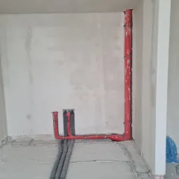 Wnętrze w trakcie prac instalacyjnych: pionowa czerwona rura przy ścianie, szare rury na podłodze i wystające kable elektryczne, surowy stan ścian.