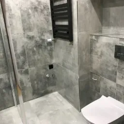 Nowoczesna łazienka z szarymi płytkami imitującymi beton, czarnym grzejnikiem drabinkowym i kabiną prysznicową ze szklanymi drzwiami.