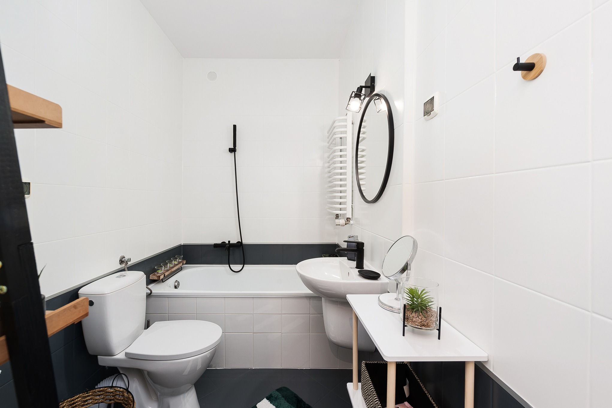 Nowoczesna, minimalistyczna łazienka z białymi płytkami, ciemnoszarą dolną częścią ścian, białą armaturą i akcentami w kolorze czarnym i drewna.
