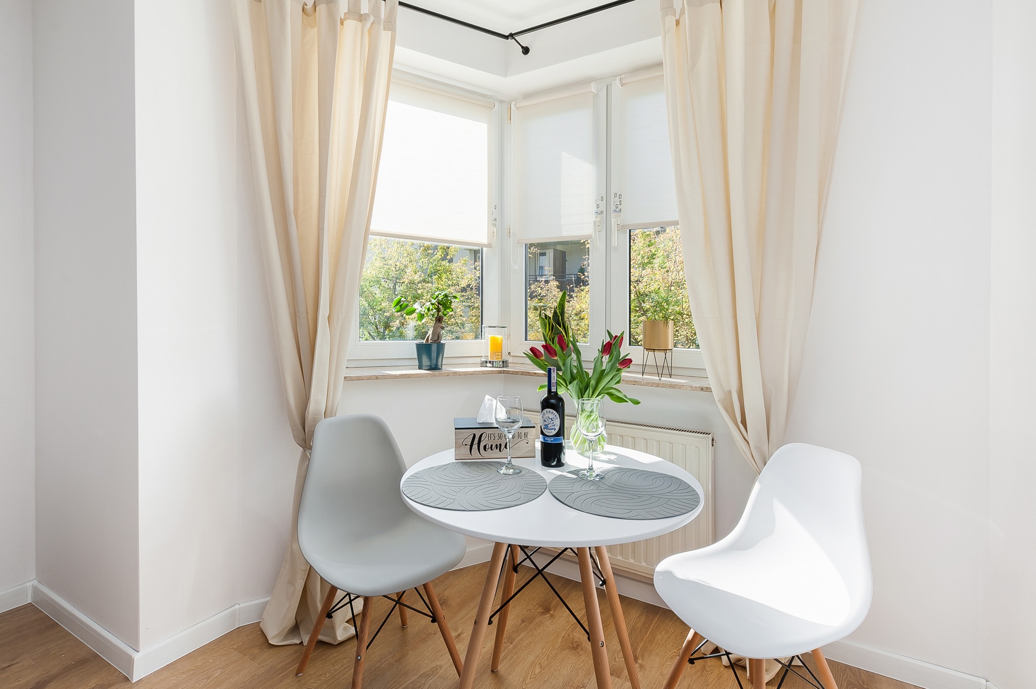 Jasne wnętrze jadalni z drewnianą podłogą, białymi ścianami, oknem z roletami i beżowymi zasłonami. Na okrągłym stole stoją tulipany w wazonie, butelka wina, dwa kieliszki, pudełko chusteczek...