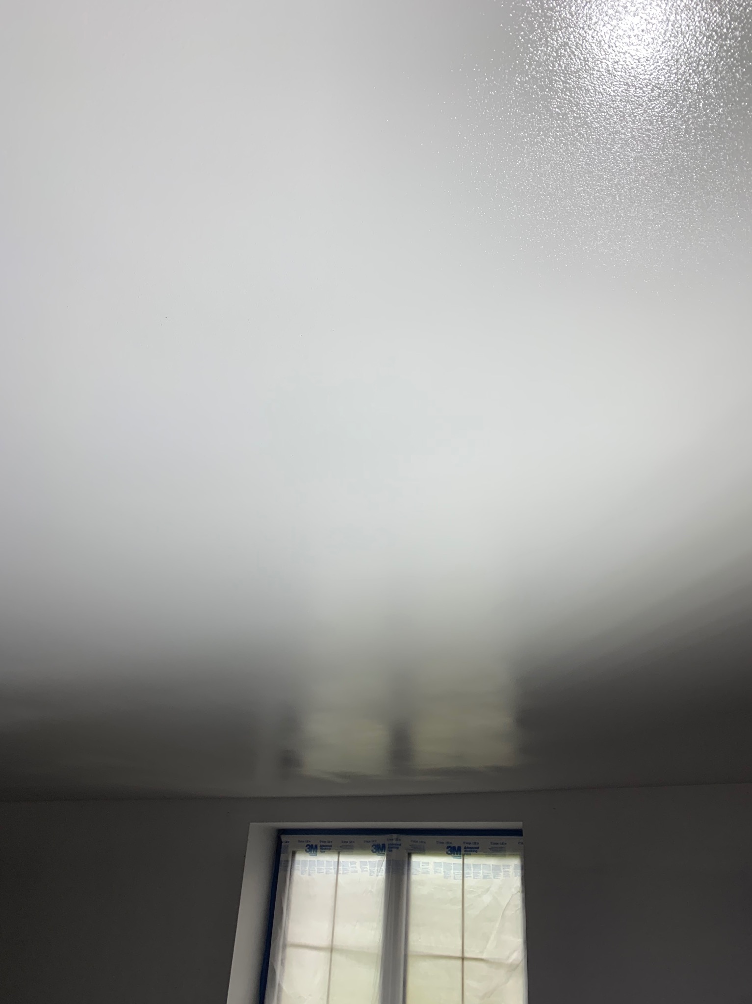 Świeżo pomalowany, biały sufit z delikatnym połyskiem, widoczne okno zabezpieczone folią malarską 3M i niepomalowane ściany.