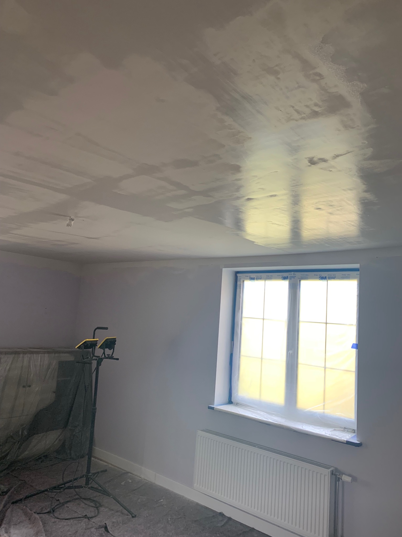 Pokój w trakcie prac wykończeniowych: sufit po nałożeniu gładzi, okno zabezpieczone folią, lampa budowlana na statywie, grzejnik. Widoczne ślady szpachlowania na suficie.