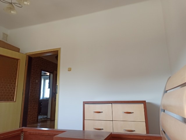 Pusta, biała ściana w pokoju po świeżym malowaniu, widoczna część mebli i drzwi do innego pomieszczenia z ceglaną ścianą.