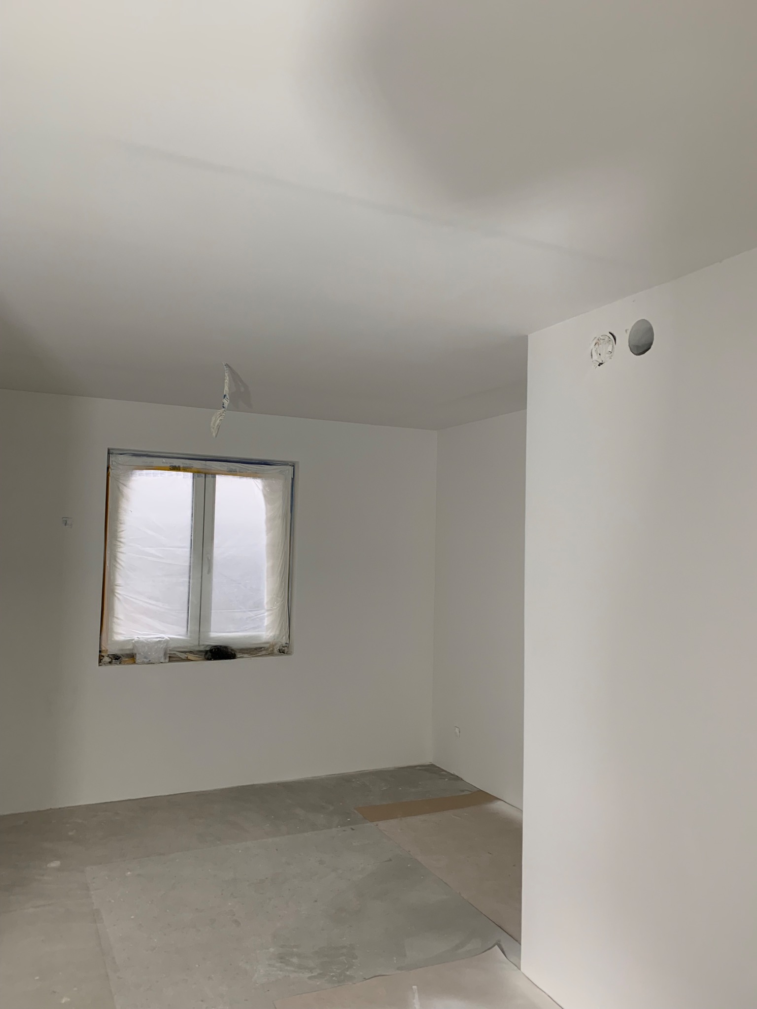 Pokój w trakcie malowania: ściany i sufit świeżo pomalowane na biało, okno zabezpieczone folią malarską, widoczne ślady po pracach na podłodze i otwory w ścianie.