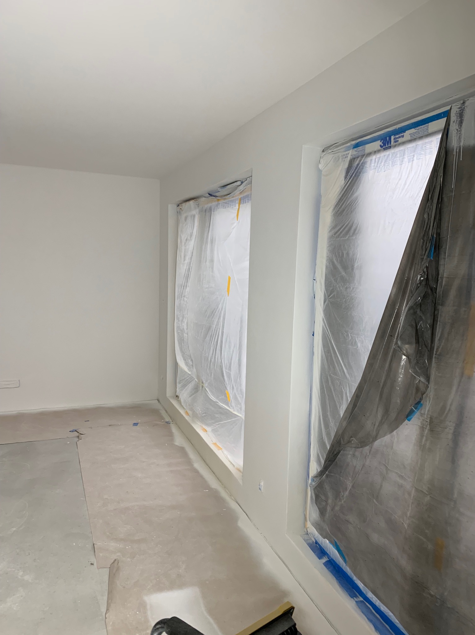 Pokój w trakcie prac malarskich, okna zabezpieczone folią malarską, ściany świeżo pomalowane na biało, podłoga częściowo zakryta papierem ochronnym.