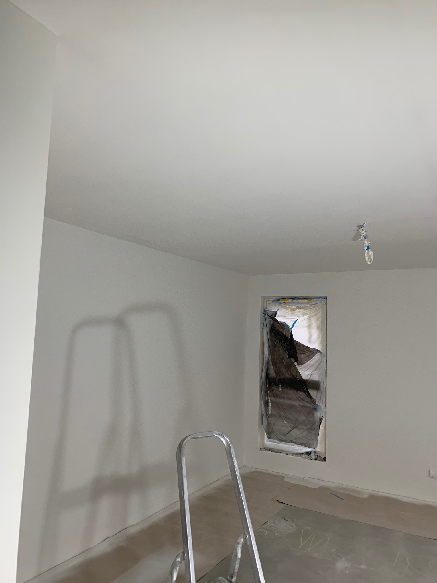 Wnętrze pokoju w trakcie odświeżania, ściany i sufit pomalowane na biało, okno zabezpieczone folią, aluminiowa drabina w rogu, widoczne ślady po pracach na podłodze.