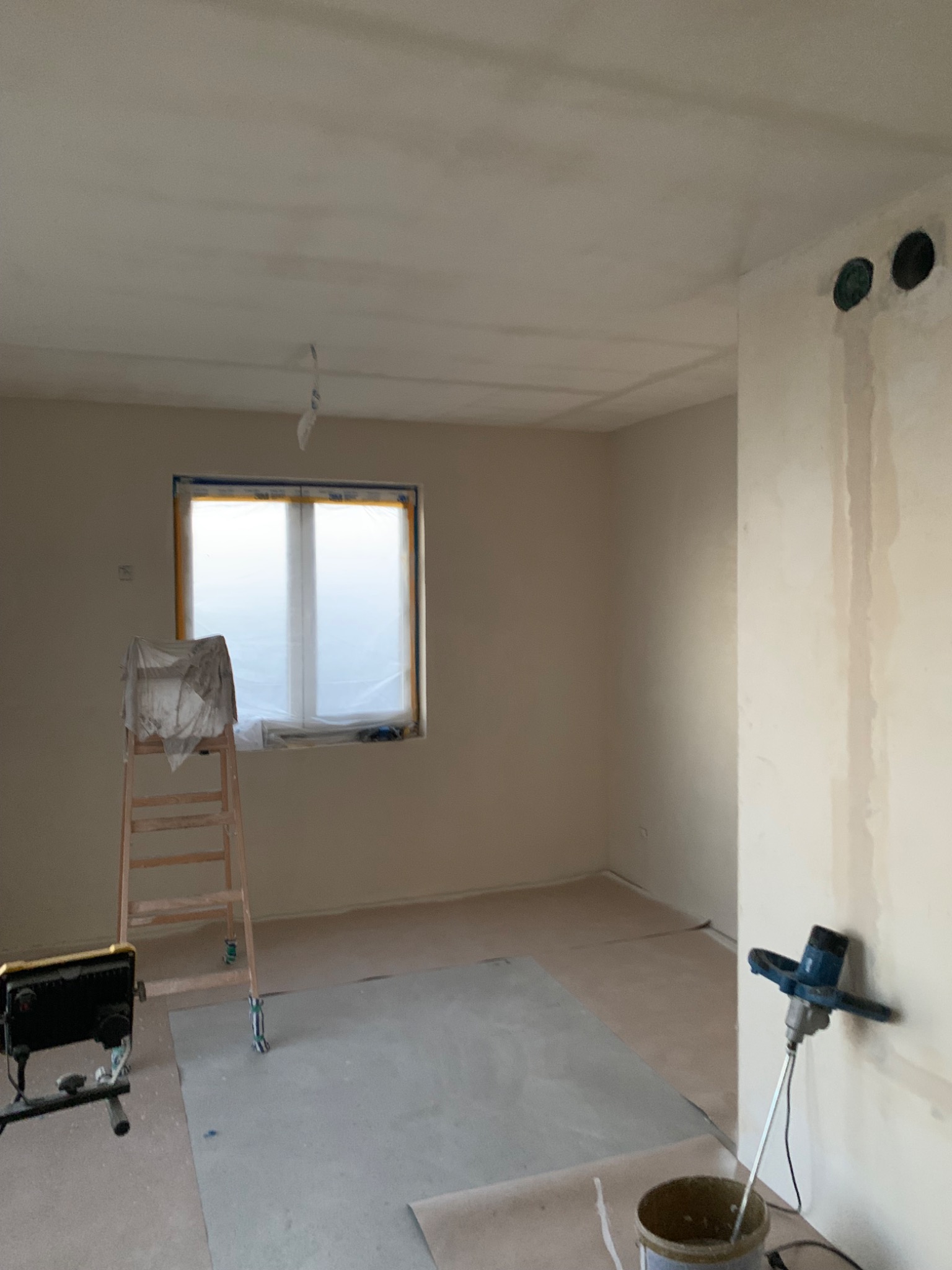Pokój w trakcie gładziowania: ściany pokryte gładzią, okno zabezpieczone folią, drabina z przykryciem, mieszadło do gładzi obok wiadra, widoczne otwory instalacyjne w ścianie.