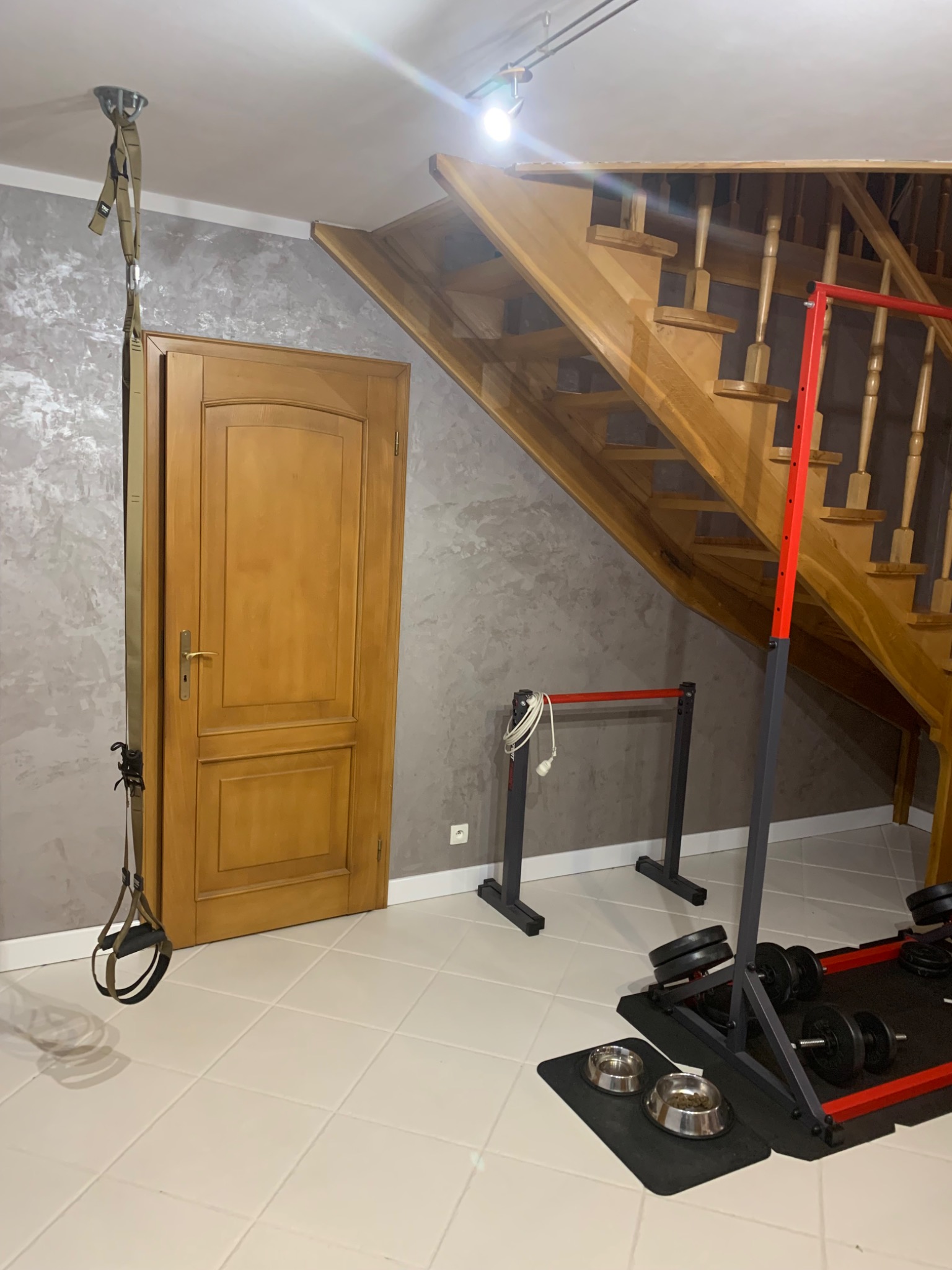 Szara ściana z efektem dekoracyjnym w domowej siłowni, widoczne drewniane schody, drzwi, sprzęt do ćwiczeń i miski dla zwierząt.