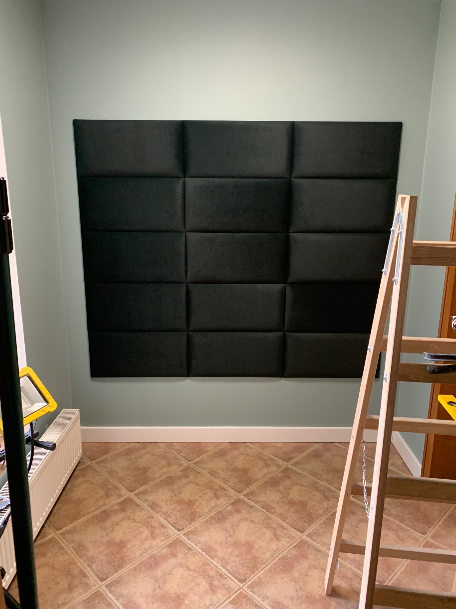 Ściana z zamontowanymi, ciemnymi panelami tapicerowanymi w układzie szesnastu kwadratów, widoczna drabina malarska i lampa halogenowa, pomieszczenie w trakcie prac wykończeniowych.