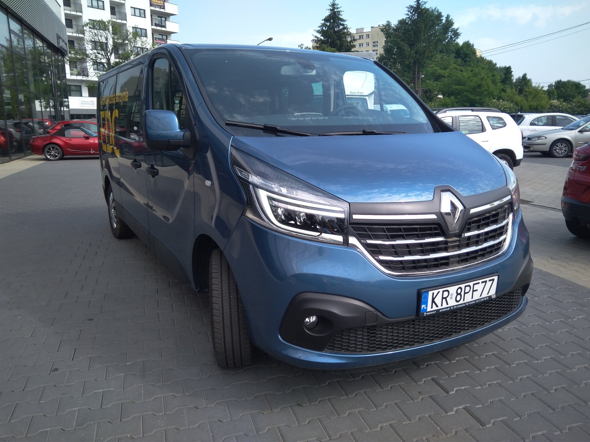 Niebieski Renault Trafic z polskimi tablicami rejestracyjnymi KR 8PF77, zaparkowany na szarym bruku z widocznym budynkiem w tle.