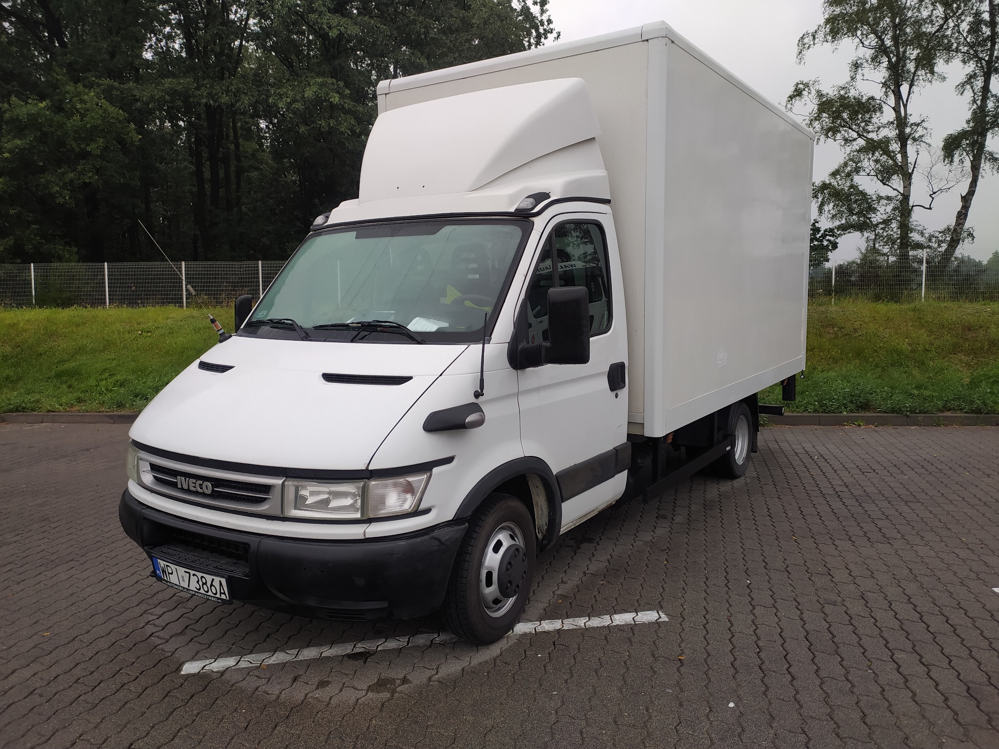 Biały samochód dostawczy Iveco z nadbudową kontenerową zaparkowany na parkingu z kostki, widoczne drzewa w tle.