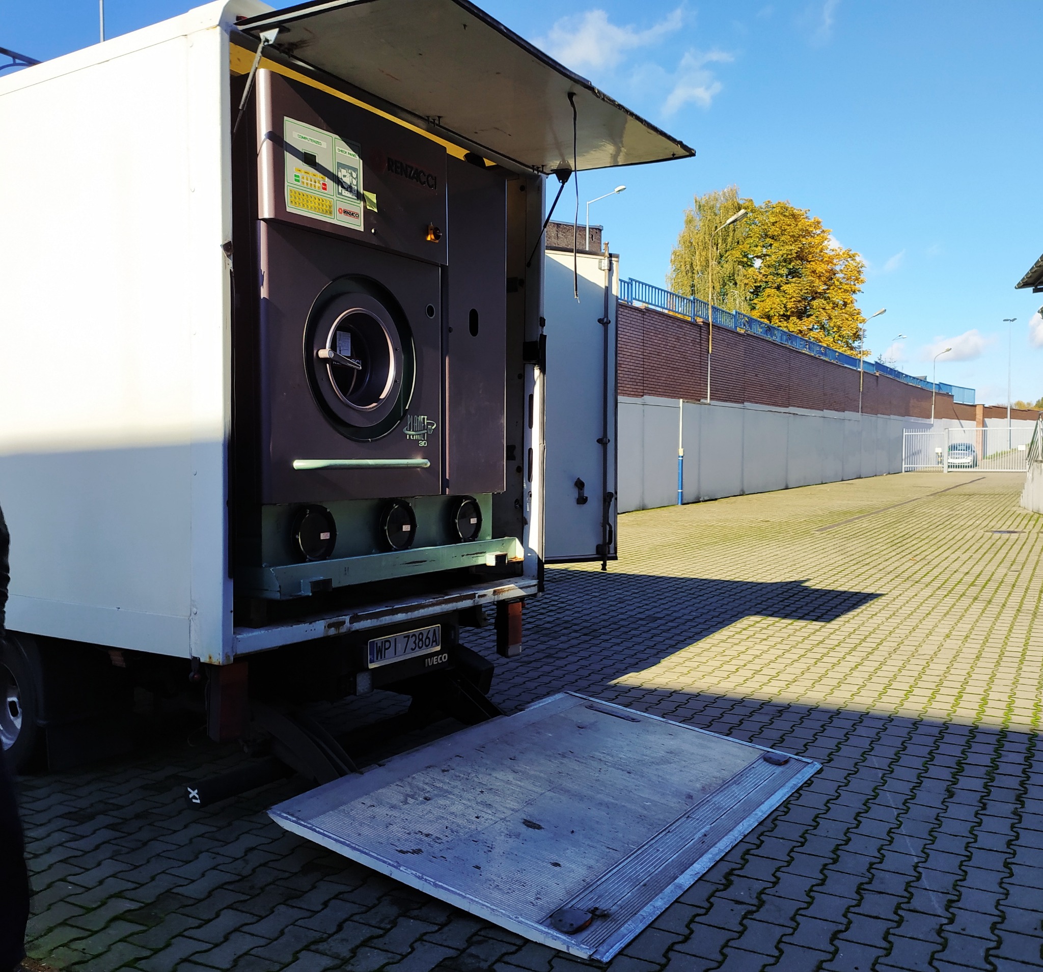 Wnętrze busa transportowego z zamontowaną specjalistyczną pralnicą przemysłową Renzacci Planet Chili 30, widoczna rampa załadunkowa i tablica rejestracyjna Iveco.