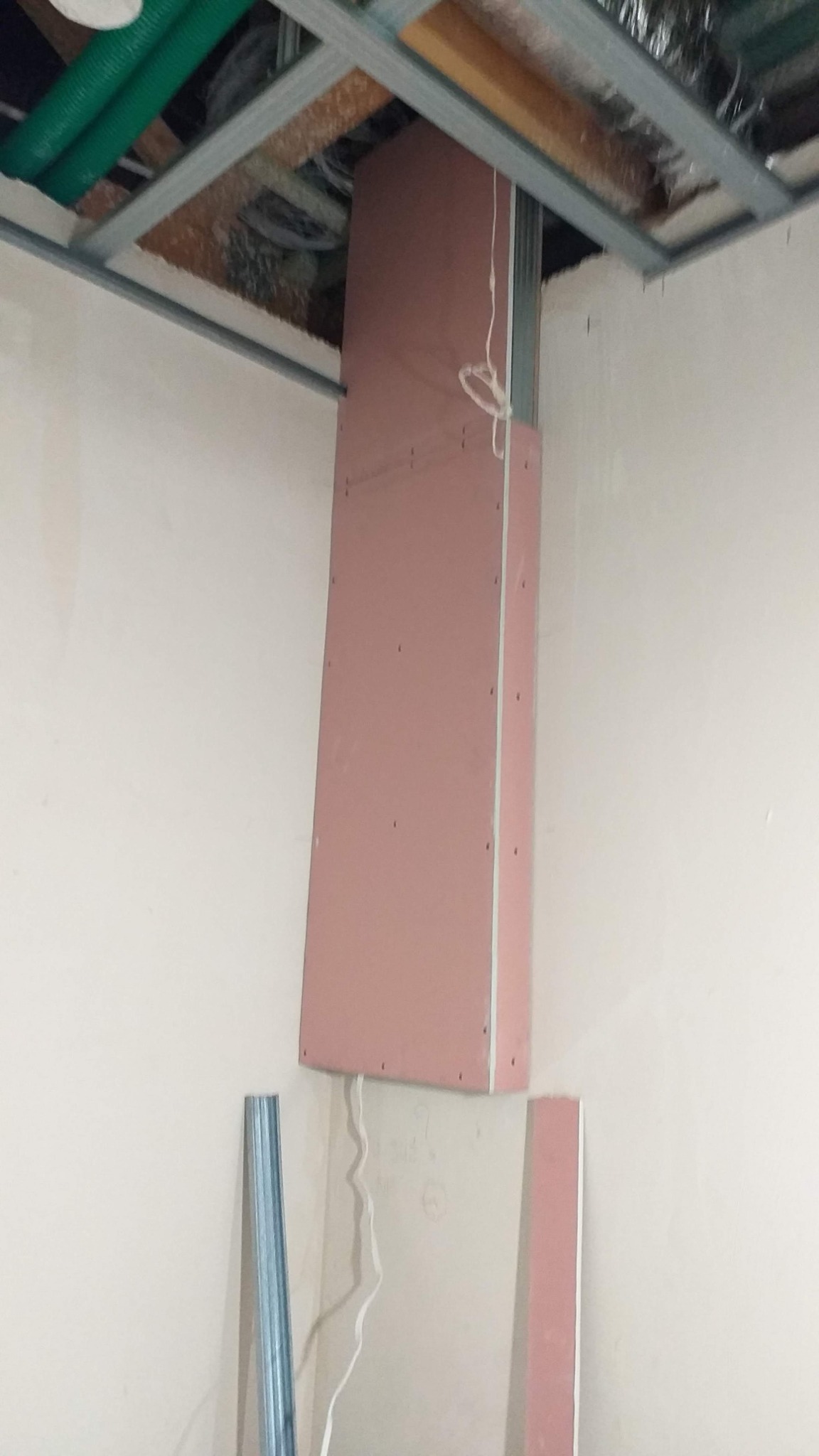 Montaż pionowej konstrukcji z różowej płyty gipsowo-kartonowej na metalowym stelażu, widoczne przewody i rura pod sufitem.