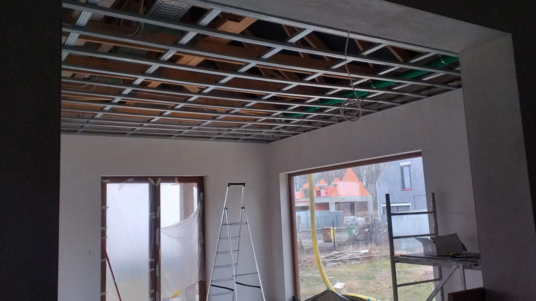 Metalowa konstrukcja sufitu podwieszanego w trakcie montażu w pomieszczeniu z oknami, widoczna drabina i elementy instalacji.
