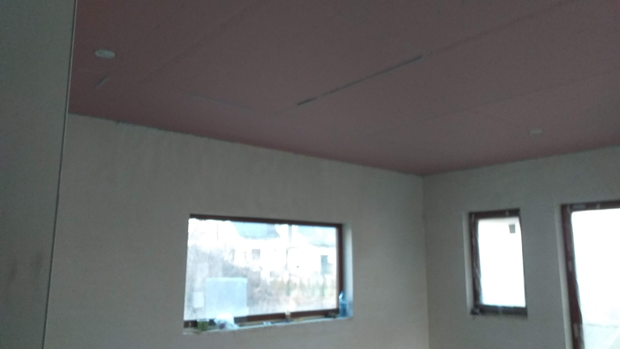 Pokój w trakcie wykańczania: sufit z różowych płyt, ściany w szarym tynku, dwa okna z brązowymi ramami.