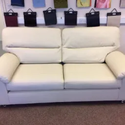 Dwuosobowa sofa z jasnej skóry ekologicznej, widoczne próbki tkanin na ścianie w tle.