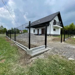 Nowoczesne ogrodzenie panelowe z podmurówką betonową w trakcie montażu, widok na dom z białymi ścianami i ciemnym dachem w tle.