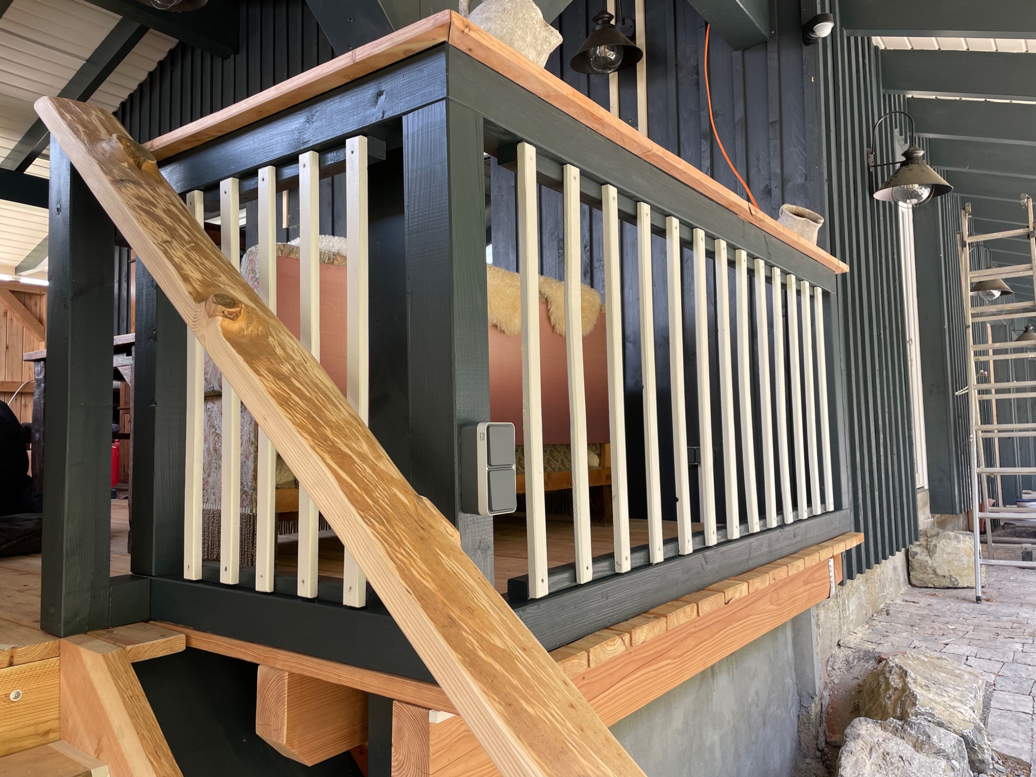 Zadaszony taras z drewnianą balustradą pomalowaną na ciemnozielono z białymi szczeblami, widoczne drewniane schody wejściowe i fragment wyposażenia wnętrza.