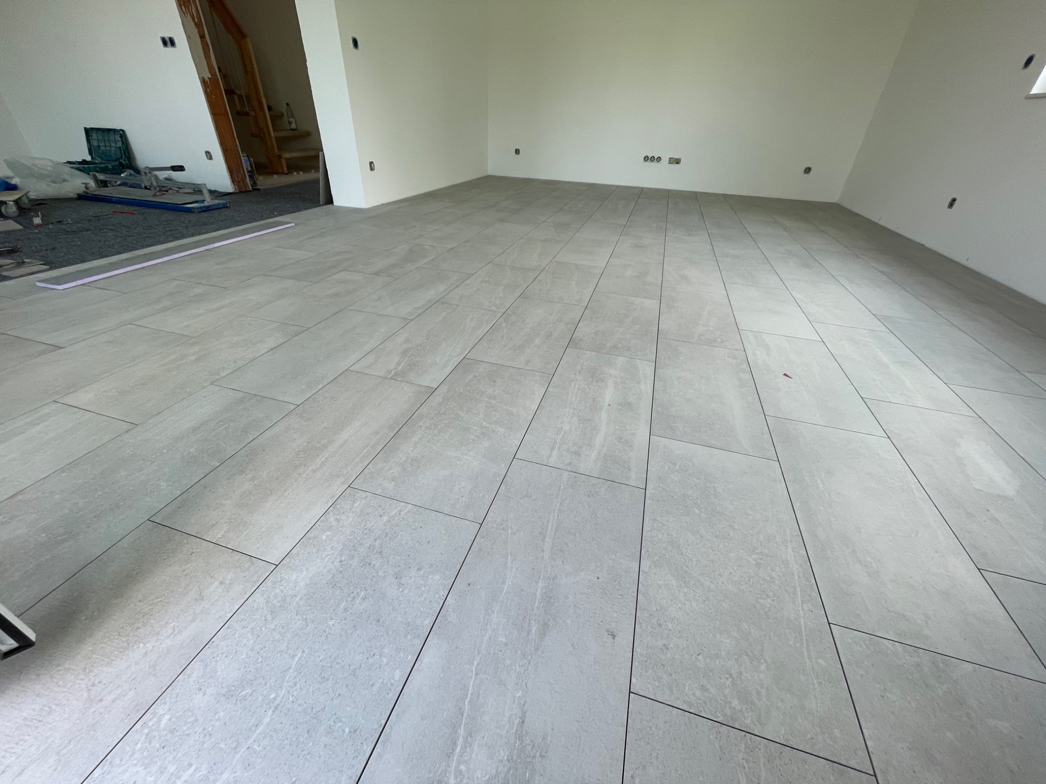 Układanie jasnoszarych, prostokątnych płytek podłogowych w dużym pomieszczeniu z białymi ścianami, widoczne narzędzia i materiały budowlane w tle.