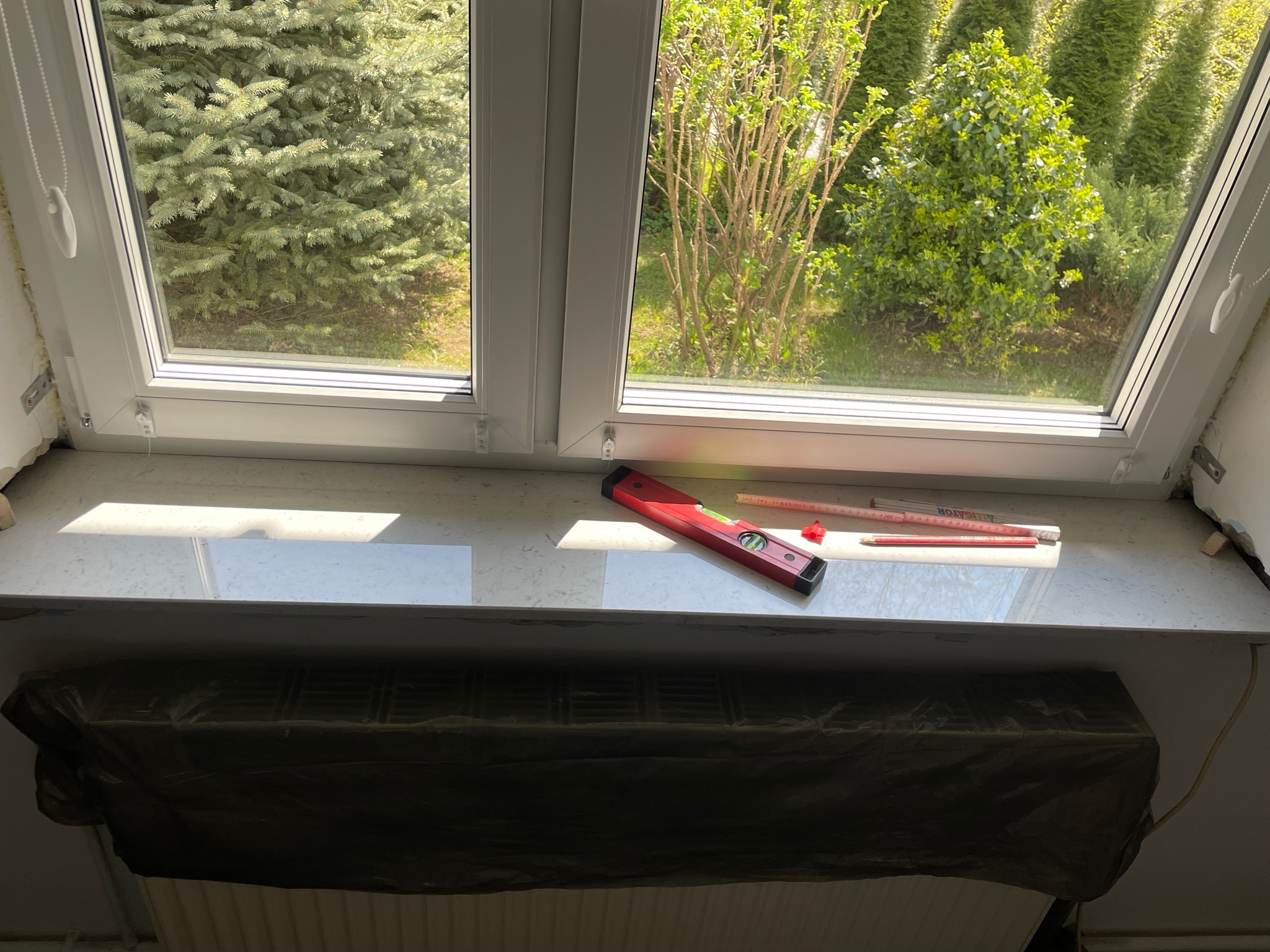 Nowy biały parapet okienny z marmurowym wzorem, poziomica, ołówek i miarka, grzejnik zabezpieczony folią malarską, widok na zieleń za oknem.