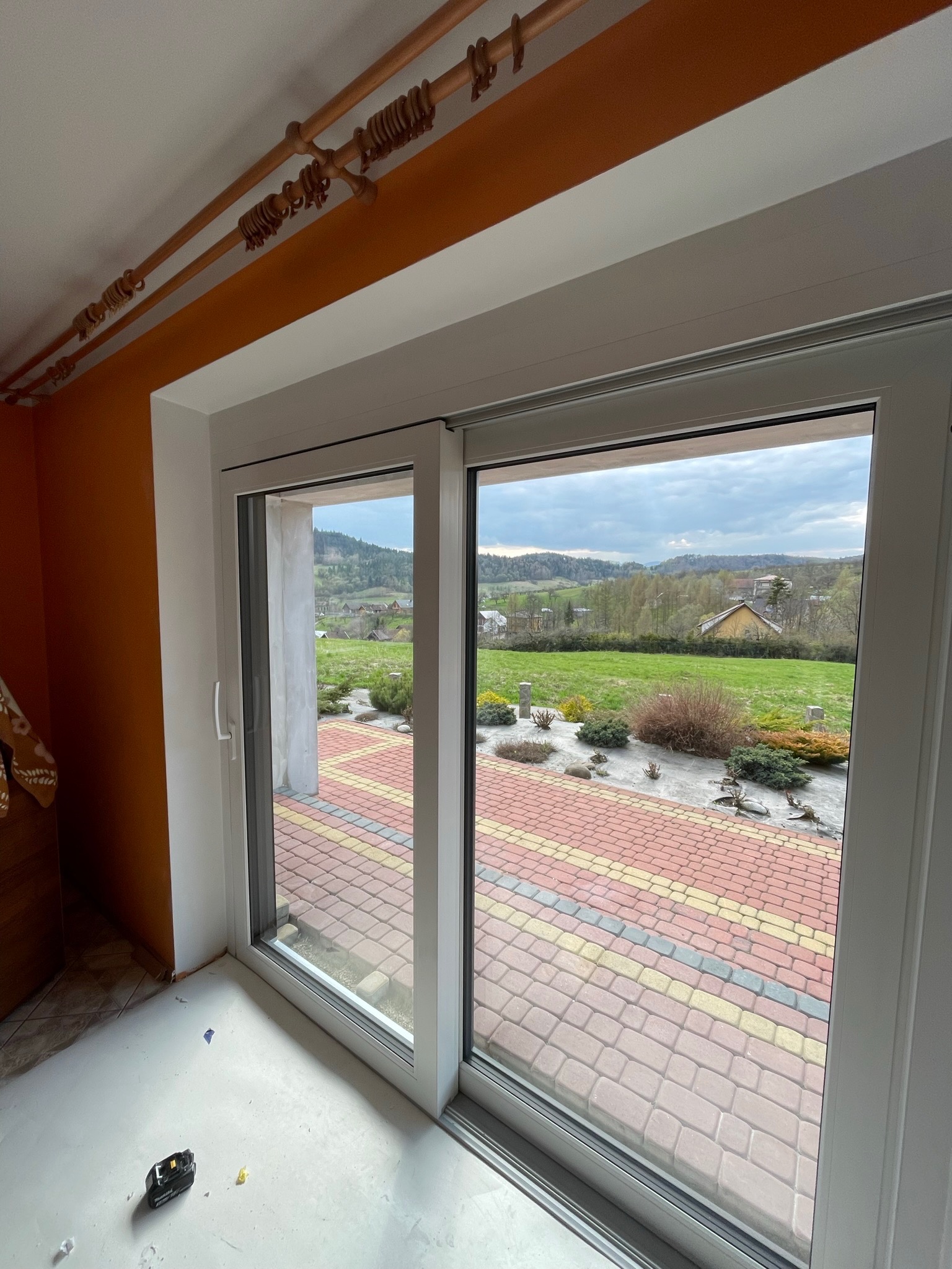 Nowo zamontowane, przesuwne drzwi balkonowe z widokiem na krajobraz wiejski i patio z kostki brukowej, widoczny poziom podłogi przed wykończeniem.