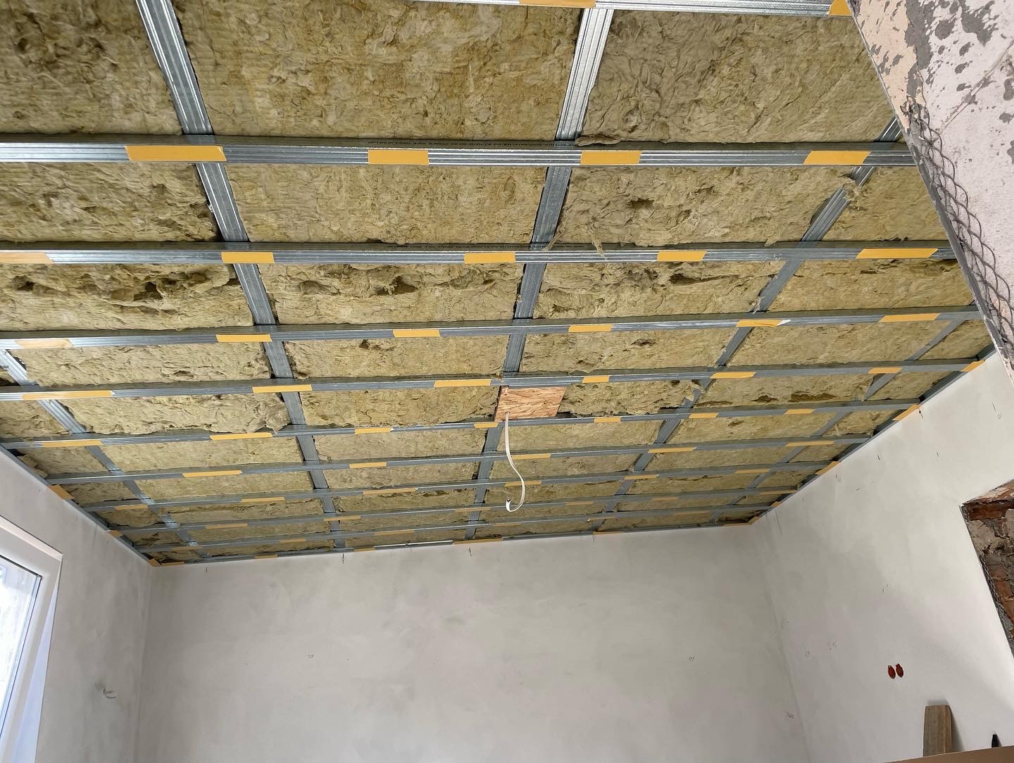 Montaż izolacji z wełny mineralnej w metalowym stelażu sufitu podwieszanego, widoczne żółte taśmy mocujące, surowy stan wykończenia wnętrza.