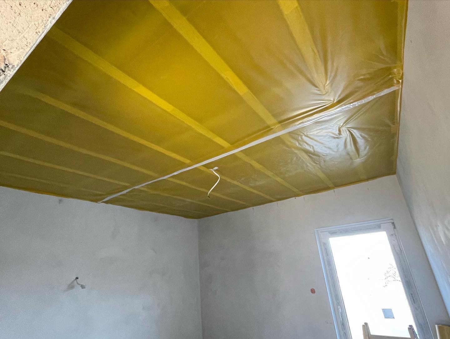 Przygotowanie sufitu do dalszych prac wykończeniowych: żółta folia ochronna przymocowana do stelaża, widoczne kable wystające z sufitu, surowe ściany w pomieszczeniu.