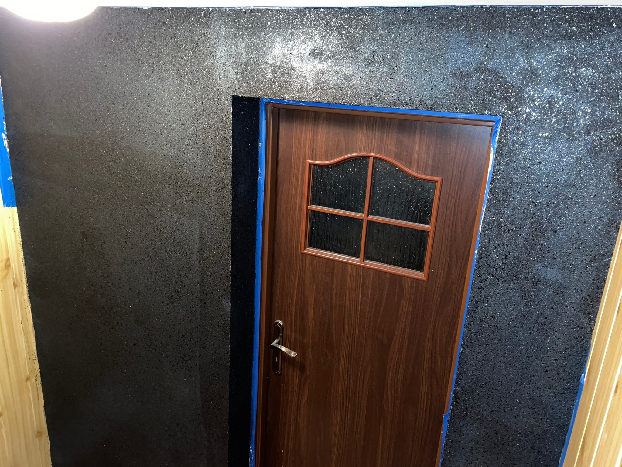 Ciemna, ziarnista ściana z efektem brokatu i brązowe drzwi z niebieską taśmą malarską na ościeżnicy, widoczne fragmenty drewnianej boazerii.
