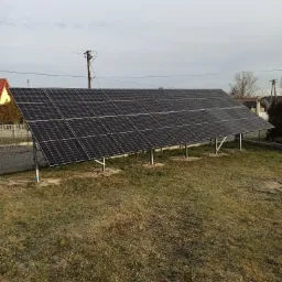 Instalacja paneli słonecznych na metalowych wspornikach na trawiastym terenie, widok z góry, domy w tle.
