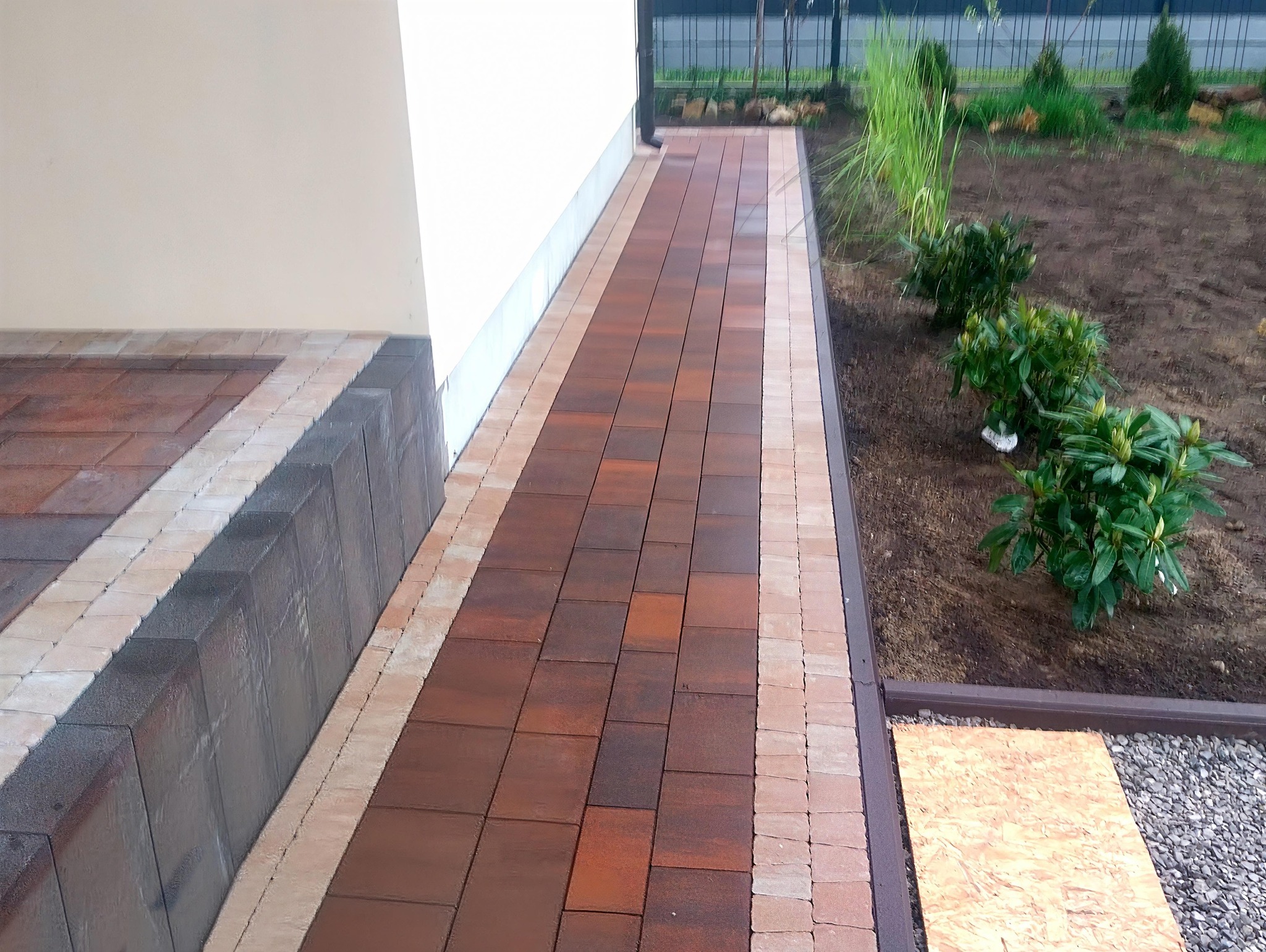 Ułożona kostka brukowa wzdłuż domu, z obrzeżami i rabatą. Brązowa kostka, jasne obrzeża. Widok z góry, lekki kąt. Świeżo posadzone rośliny.